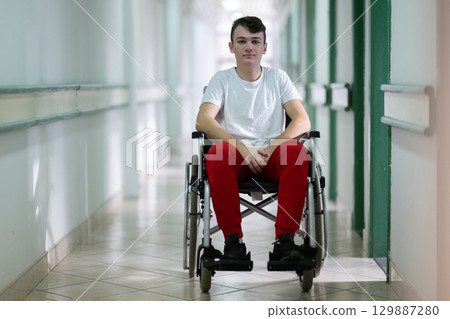 Young teenage boy smiling while sitting in a wheelchair in a hospital hallway. Rehabilitation and positivity concept. Young teenage boy smiling while sitting in a wheelchair in a hospital hallway. Rehabilitation and positivity concept. 129887280