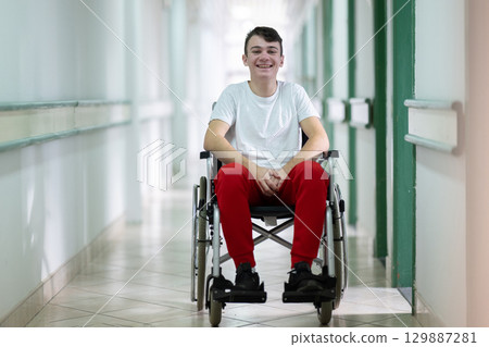 Young teenage boy smiling while sitting in a wheelchair in a hospital hallway. Rehabilitation and positivity concept. Young teenage boy smiling while sitting in a wheelchair in a hospital hallway. Rehabilitation and positivity concept. 129887281