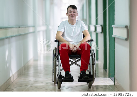 Young teenage boy smiling while sitting in a wheelchair in a hospital hallway. Rehabilitation and positivity concept. 129887283