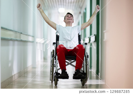 Joyful Young Man in Wheelchair.Teenage boy celebrating in wheelchair with raised arms in hospital hallway. Positivity and recovery. Joyful Young Man in Wheelchair.Teenage boy celebrating in wheelchair with raised arms in hospital hallway. Positivity and recovery. 129887285