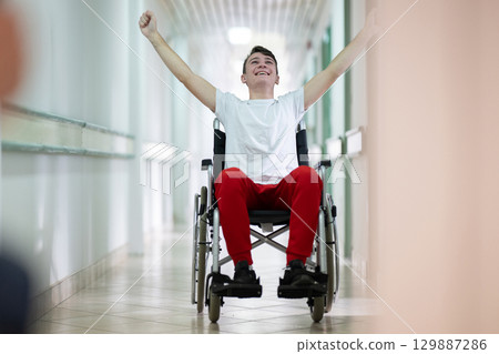 Joyful Young Man in Wheelchair.Teenage boy celebrating in wheelchair with raised arms in hospital hallway. Positivity and recovery. Joyful Young Man in Wheelchair.Teenage boy celebrating in wheelchair with raised arms in hospital hallway. Positivity and recovery. 129887286