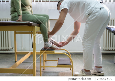Physical Therapist Assisting Elderly Patient.Physiotherapist helping an elderly woman with leg movement on a step platform during a rehabilitation session. 129887399
