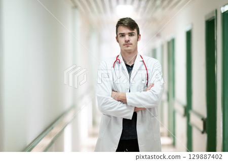 Doctor Standing in Hospital Hallway.Young male doctor standing confidently in a hospital corridor with a stethoscope. Medical professionalism concept. Doctor Standing in Hospital Hallway.Young male doctor standing confidently in a hospital corridor with a stethoscope. Medical professionalism concept. 129887402