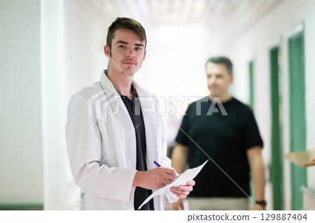 Doctor Reading Documents in Hospital.Young male doctor walking in hospital hallway while reading patient documentation. Clinical work environment. 129887404