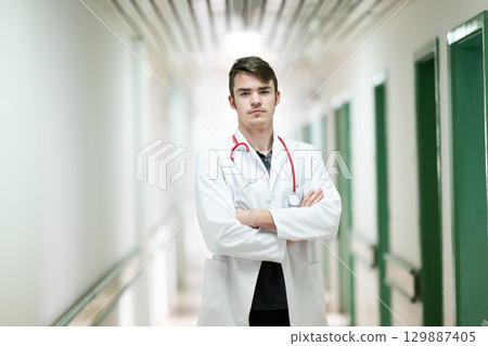 Confident Young Doctor Portrait.Portrait of a young doctor in white coat with arms crossed, standing in a hospital hallway. Confident Young Doctor Portrait.Portrait of a young doctor in white coat with arms crossed, standing in a hospital hallway. 129887405