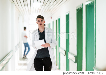Doctor Reading Documents in Hospital.Young male doctor walking in hospital hallway while reading patient documentation. Clinical work environment. 129887412