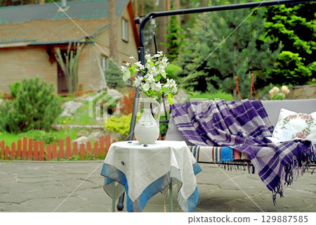 Apple blossoms on a table in front of a sofa swing in the courtyard of a country house. Apple blossoms on a table in front of a sofa swing in the courtyard of a country house. 129887585