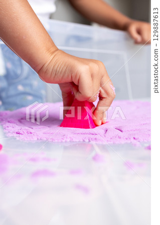 Child hand pressing pink cone into purple sand for creative play activity 129887613