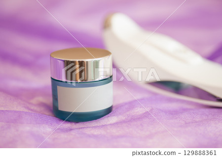 Small plastic jar of face cream or scrub on violet background, close up, copy space. Concept of cosmetic product, skin care, dermatology, wellness and cosmetology 129888361
