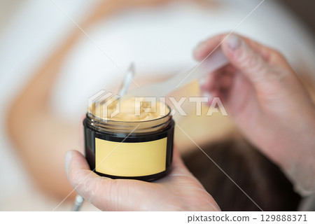 Brown half transparent plastic container with facial mask in female hands, copy space. Professional cosmetologist taking mask to apply in it on clients face Brown half transparent plastic container with facial mask in female hands, copy space. Professional cosmetologist taking mask to apply in it on clients face 129888371
