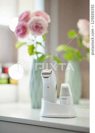 Devices for ultrasonic facial cleansing and massage standing on the table, close up. Mirror and beautiful rose flowers on the background in the beauty salon 129888392