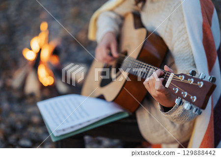 Close up of a girl guitarist practicing on nature near the camp fire. Warm evening shot of a girl playing the guitar with notebook, composing new song near bonfire 129888442