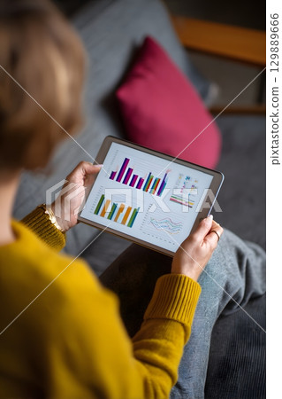 Woman sitting on sofa using digital tablet for virtual meeting with charts 129889666