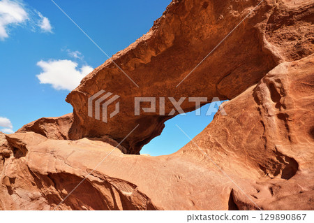 Little arc or small rock window formation in Wadi Rum desert, bright sun shines on red dust and rocks, blue sky above 129890867