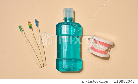 Set of oral hygiene products and items, including three toothbrushes, a bottle of mouthwash and a model of a jaw, beige background. 129892045