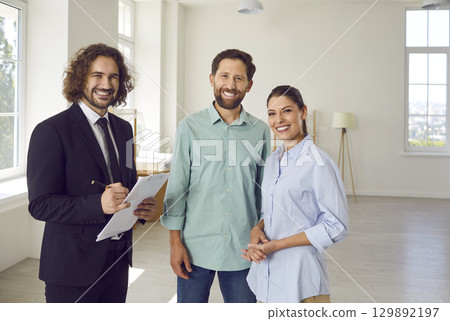 Young man realtor standing with happy couple looking at camera in new house. Young man realtor standing with happy couple looking at camera in new house. 129892197