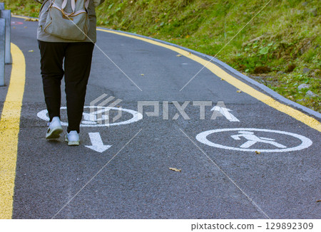 A person walks along a pedestrian path, following the marked direction on the ground. A person walks on a path marked for pedestrians, with directional signs and a backpack, on a sunny day. A person walks along a pedestrian path, following the marked direction on the ground. A person walks on a path marked for pedestrians, with directional signs and a backpack, on a sunny day. 129892309