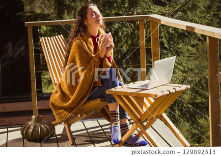 Relaxing woman on a wooden balcony enjoying coffee and working on laptop in the forest setting. A calm girl enjoys tea drink and sunlight on a wooden terrace, wrapped in a cozy blanket enjoying warmth Relaxing woman on a wooden balcony enjoying coffee and working on laptop in the forest setting. A calm girl enjoys tea drink and sunlight on a wooden terrace, wrapped in a cozy blanket enjoying warmth 129892313