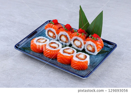 Sushi rolls wrapped in salmon and filled with tempura shrimp, topped with red tobiko and microgreens, served on a decorative blue plate. Sushi rolls wrapped in salmon and filled with tempura shrimp, topped with red tobiko and microgreens, served on a decorative blue plate. 129893056