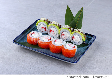 Colorful sushi rolls with salmon, avocado, shrimp, cream cheese, and roe, topped with sauce and crunchy bits, served on a blue rectangular plate. Colorful sushi rolls with salmon, avocado, shrimp, cream cheese, and roe, topped with sauce and crunchy bits, served on a blue rectangular plate. 129893058