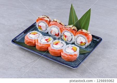 Salmon-wrapped sushi rolls filled with shrimp, mango, cream cheese, and roe, topped with almond slices and served on a decorative plate Salmon-wrapped sushi rolls filled with shrimp, mango, cream cheese, and roe, topped with almond slices and served on a decorative plate 129893062