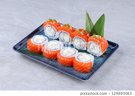 Salmon sushi rolls with shrimp, cream cheese, and fruit filling, garnished with microgreens and crunchy toppings on a blue plate. 129893063