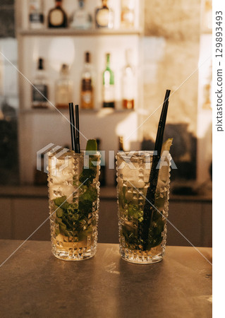 Two beautifully crafted mojitos sit on a bar ready to be enjoyed. Fresh mint, lime and straws in a refreshing cocktail. Cold summer drink. Two beautifully crafted mojitos sit on a bar ready to be enjoyed. Fresh mint, lime and straws in a refreshing cocktail. Cold summer drink. 129893493