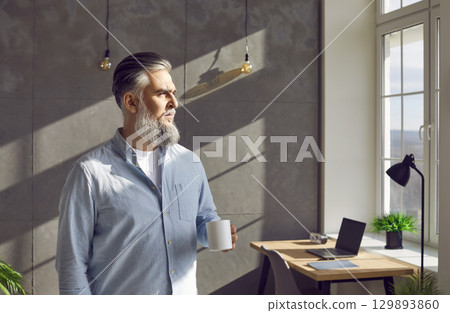 Mature business man standing by the window near his workplace at office with cup of coffee. Mature business man standing by the window near his workplace at office with cup of coffee. 129893860