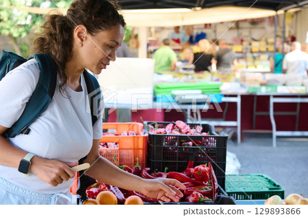 A woman explores a vibrant outdoor market, handpicking fresh produce like fruits and vegetables. The scene reflects a healthy lifestyle, eco-consciousness, and the charm of local markets. 129893866