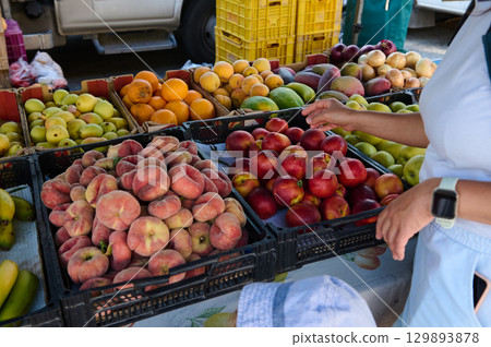 Colorful selection of fresh fruits and vegetables on an outdoor market stand, with a customer choosing produce. Perfect for promoting healthy living, vegan and vegetarian diets, and organic choices. Colorful selection of fresh fruits and vegetables on an outdoor market stand, with a customer choosing produce. Perfect for promoting healthy living, vegan and vegetarian diets, and organic choices. 129893878