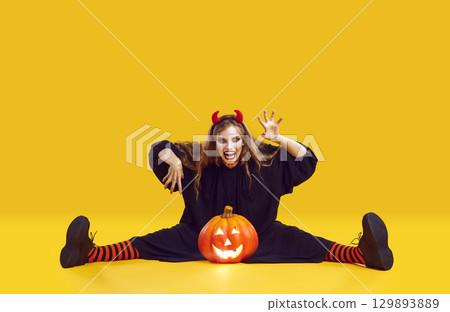Frightening crazy woman with Halloween makeup sitting with pumpkin on orange background. 129893889