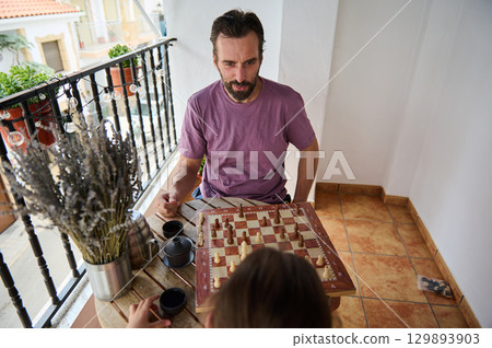 A thoughtful father plays chess with his daughter on a cozy sunlit balcony. The scene captures a serene moment with tea, outdoor ambiance, and quality family interaction representing relaxation and 129893903