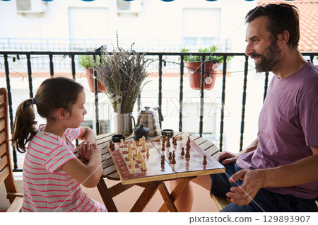 A father and daughter enjoy a friendly game of chess on a balcony decorated with potted plants. The setting is warm and inviting, with a focus on bonding and mindful moments. 129893907