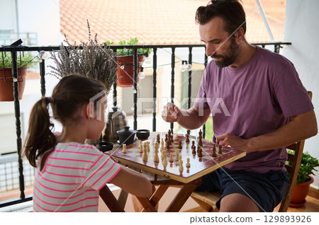 A father and his young daughter share a quality moment playing chess on a sunny balcony, surrounded by potted plants and enjoying the peaceful ambiance. A father and his young daughter share a quality moment playing chess on a sunny balcony, surrounded by potted plants and enjoying the peaceful ambiance. 129893926