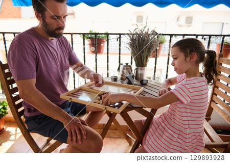 A tranquil moment of a father and his young daughter playing backgammon on a balcony table. Natural light highlights the bond in this casual setting, emphasizing family connection and leisure. A tranquil moment of a father and his young daughter playing backgammon on a balcony table. Natural light highlights the bond in this casual setting, emphasizing family connection and leisure. 129893928