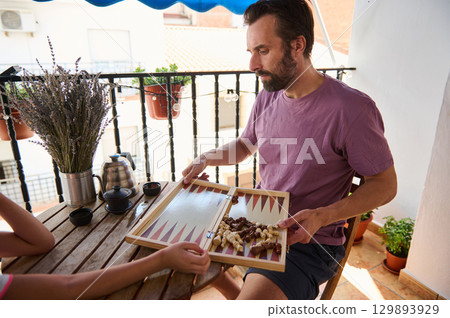 A man sits on a sunny balcony engaging with a backgammon game. Decorated with plants and tea set, the serene setting invites relaxation and quality time. 129893929