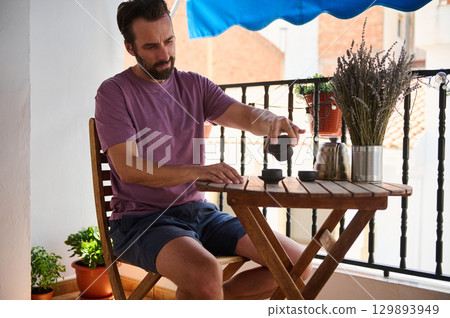 A man sits outdoors on a balcony with wooden furniture, preparing tea. A comfortable and serene setting is complemented by potted plants and a table arrangement, showcasing a calming and peaceful 129893949