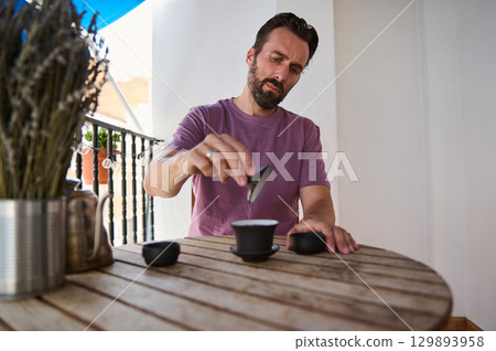 A man sitting at a wooden table on a bright balcony performs a tea ceremony. He appears tranquil and focused, creating a serene and harmonious setting with natural decor elements around him. A man sitting at a wooden table on a bright balcony performs a tea ceremony. He appears tranquil and focused, creating a serene and harmonious setting with natural decor elements around him. 129893958