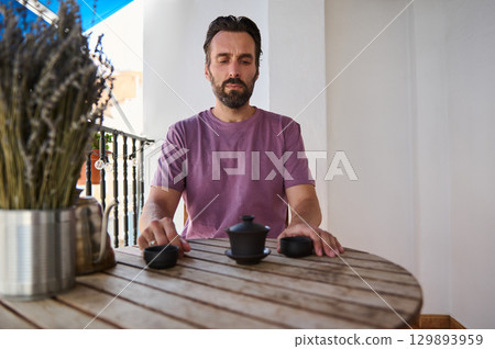 Serene outdoor moment of a man contemplating life while enjoying tea on a sunny balcony, surrounded by natural elements. Peaceful scene to reflect solitude, simplicity, and lifestyle relaxation. 129893959