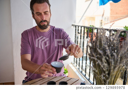 A man performs a tea ceremony on a sunny balcony decorated with plants and lavender. This serene scene highlights personal relaxation, traditional practices, and the enjoyment of a quiet moment. A man performs a tea ceremony on a sunny balcony decorated with plants and lavender. This serene scene highlights personal relaxation, traditional practices, and the enjoyment of a quiet moment. 129893978