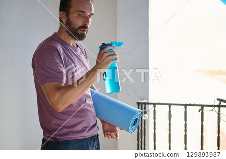 Bearded man on a balcony holding a yoga mat and water bottle, ready for a fitness session. The bright ambiance highlights a focus on health, wellness, and mindfulness in a cozy home setting. 129893987