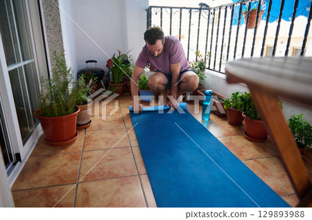 A young man in casual athletic wear organizing his yoga mat after exercising on a balcony adorned with lush potted plants, highlighting personal health and outdoor wellness. 129893988