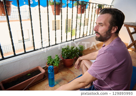 A bearded man relaxes on his balcony, surrounded by potted plants and enjoying a serene moment seated beside a yoga mat. 129893993