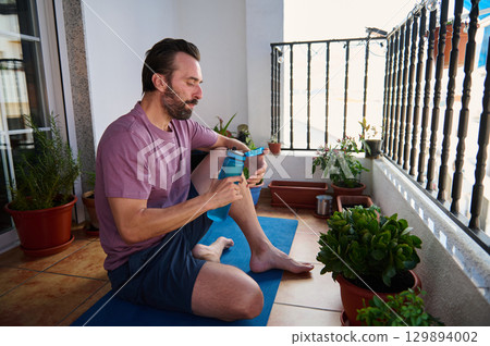 Man sitting on balcony yoga mat surrounded by plants, enjoying tea after exercise. The serene atmosphere highlights relaxation and a healthy lifestyle in an outdoor urban sanctuary. Man sitting on balcony yoga mat surrounded by plants, enjoying tea after exercise. The serene atmosphere highlights relaxation and a healthy lifestyle in an outdoor urban sanctuary. 129894002