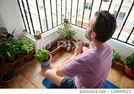 A man sits cross-legged, meditating on a yoga mat amidst potted plants on a cozy balcony. The scene embodies tranquility and mindfulness, highlighting a personal fitness and relaxation routine. 129894025