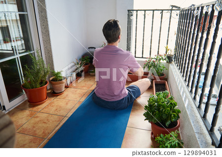 A young man meditates on his balcony, sitting on a yoga mat surrounded by potted plants. The serene environment exudes relaxation and mindfulness, highlighting the bond with nature in urban settings. A young man meditates on his balcony, sitting on a yoga mat surrounded by potted plants. The serene environment exudes relaxation and mindfulness, highlighting the bond with nature in urban settings. 129894031