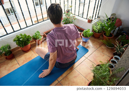 A man performs a yoga pose on his balcony surrounded by potted plants, embracing peace. 129894033