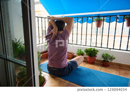 A man sits on a yoga mat on a residential balcony, surrounded by plants, performing a stretching exercise. The tranquil urban setting provides a peaceful environment for relaxation and fitness. 129894034