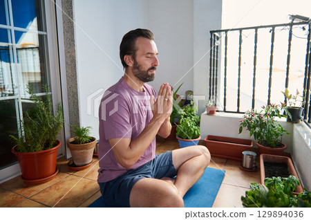 Man meditating on a balcony surrounded by potted plants, sitting on a yoga mat. The serene atmosphere reflects mindfulness, relaxation, and a healthy lifestyle in an urban outdoor setting. Man meditating on a balcony surrounded by potted plants, sitting on a yoga mat. The serene atmosphere reflects mindfulness, relaxation, and a healthy lifestyle in an urban outdoor setting. 129894036