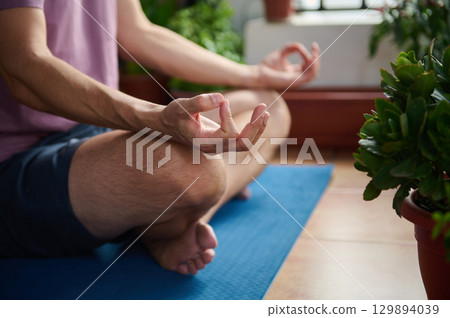 A man sits cross-legged on a yoga mat indoors, meditating surrounded by potted plants, creating a peaceful and serene atmosphere. A man sits cross-legged on a yoga mat indoors, meditating surrounded by potted plants, creating a peaceful and serene atmosphere. 129894039